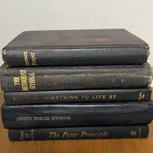 May include: A collection of five antique books with black covers. The titles visible are "Common Prayer," "The Methodist Hymnal," "Kopplin - Something to Live By," "Armed Forces Hymnal," and "The Peter Principle." The books are stacked.