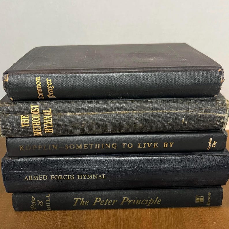 May include: A collection of five antique books with black covers. The titles visible are "Common Prayer," "The Methodist Hymnal," "Kopplin - Something to Live By," "Armed Forces Hymnal," and "The Peter Principle." The books are stacked.