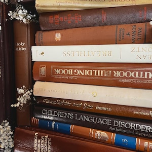 Op de afbeelding: Een stapel vintage boeken in verschillende kleuren, waaronder bruin, beige en donker bordeauxrood. Titels zijn onder meer "Holy Bible", "Breathless" en "Children's Language Disorders". Sommige boeken hebben gouden letters en decoratieve accenten. Witte bloemen zijn aan de zijkant geplaatst.