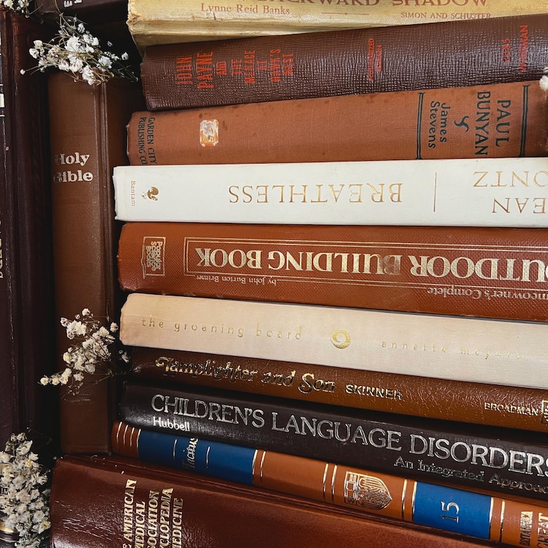 Op de afbeelding: Een stapel vintage boeken in verschillende kleuren, waaronder bruin, beige en donker bordeauxrood. Titels zijn onder meer "Holy Bible", "Breathless" en "Children's Language Disorders". Sommige boeken hebben gouden letters en decoratieve accenten. Witte bloemen zijn aan de zijkant geplaatst.