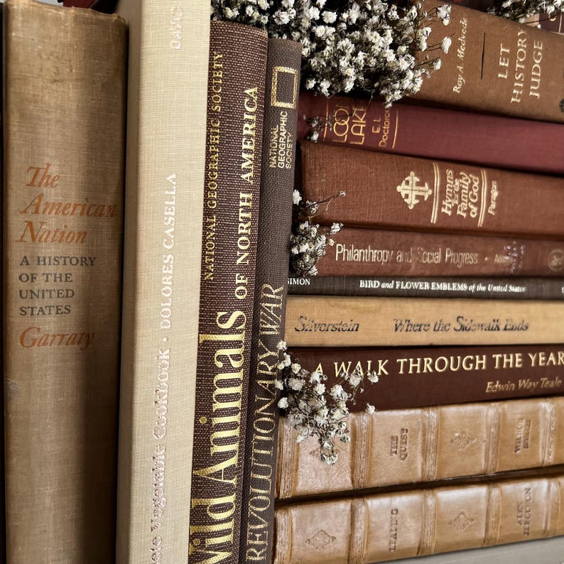Op de afbeelding: Een verzameling vintage boeken in bruine en beige tinten, met gouden letters. Titels zijn onder meer "Wild Animals of North America" en "Revolutionary War". Kleine witte bloemen zijn tussen de boeken geplaatst.