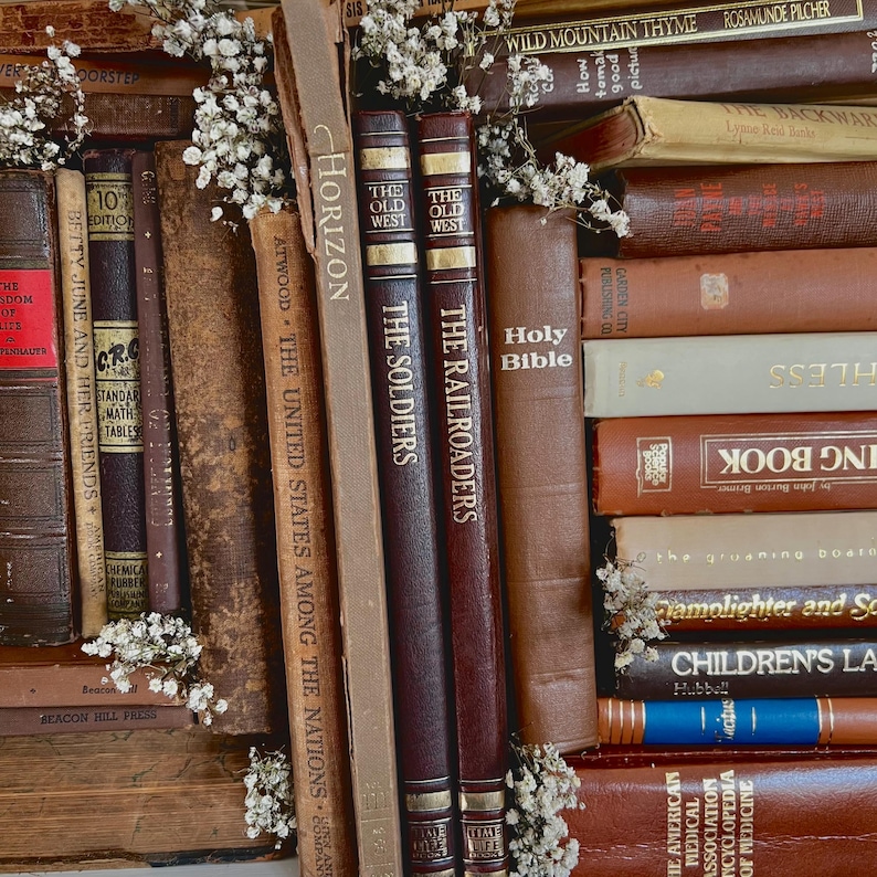 Op de afbeelding: Een verzameling vintage boeken in bruine en bordeauxrode tinten, met gouden letters. Titels zoals "The Soldiers" en "Holy Bible" zijn zichtbaar. Kleine witte bloemen zijn tussen de boeken geplaatst.