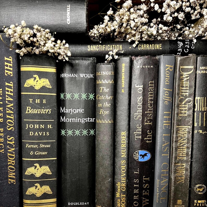May include: A selection of antique books with black covers and gold lettering. Titles include "The Bouviers," "Marjorie Morningstar," and "The Catcher in the Rye." Small white flowers are arranged amongst the books, creating a classic look.