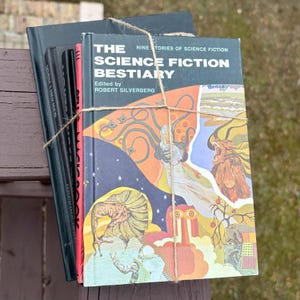 May include: A collection of vintage science fiction books bound with string. The top book, "The Science Fiction Bestiary," displays vibrant illustrations of mythical beings. Other books are black with red spines.