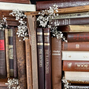 Op de afbeelding: Een verzameling vintage boeken in verschillende bruin- en beigetinten, met titels als "The Soldiers" en "Holy Bible". Kleine witte bloemen zijn tussen de boeken geplaatst, wat een rustieke en vintage uitstraling cre&euml;ert. De boeken zijn op elkaar gestapeld.