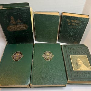 May include: A collection of antique books with dark green covers. Titles include "The Popular Educator," "The Ministry of Music," and "The One Woman." Some books feature gold lettering and embossed designs. The books are arranged on a light surface.