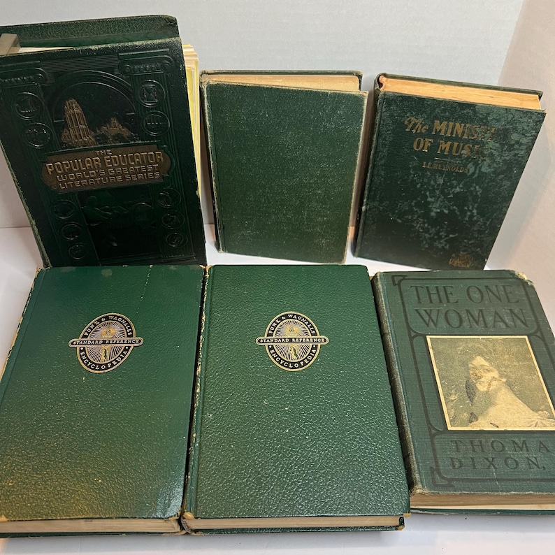May include: A collection of antique books with dark green covers. Titles include "The Popular Educator," "The Ministry of Music," and "The One Woman." Some books feature gold lettering and embossed designs. The books are arranged on a light surface.
