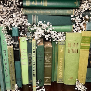 May include: A collection of vintage books in various shades of green and yellow, arranged with white baby's breath flowers. Titles include "Old Charlie," "Human Action," and "Easy German."