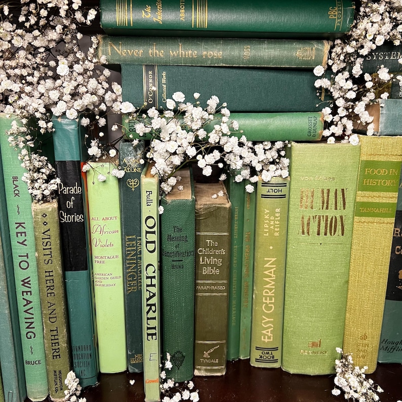 May include: A collection of vintage books in various shades of green and yellow, arranged with white baby's breath flowers. Titles include "Old Charlie," "Human Action," and "Easy German."