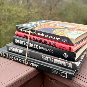 May include: A pile of old books bound with string. Titles visible are "The Science Fiction Bestiary", "Bells Magic Book", "Lightsource", "Frontline of Discovery", and "Hacking Innovation". The books are in various colours, including red, black, and blue.