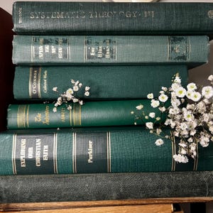 May include: A stack of vintage books in varying shades of green, with gold lettering and accents. Titles include "Systematic Theology", "Exploring Our Christian Faith", and "The Inheritors". Delicate white flowers are nestled among the books.