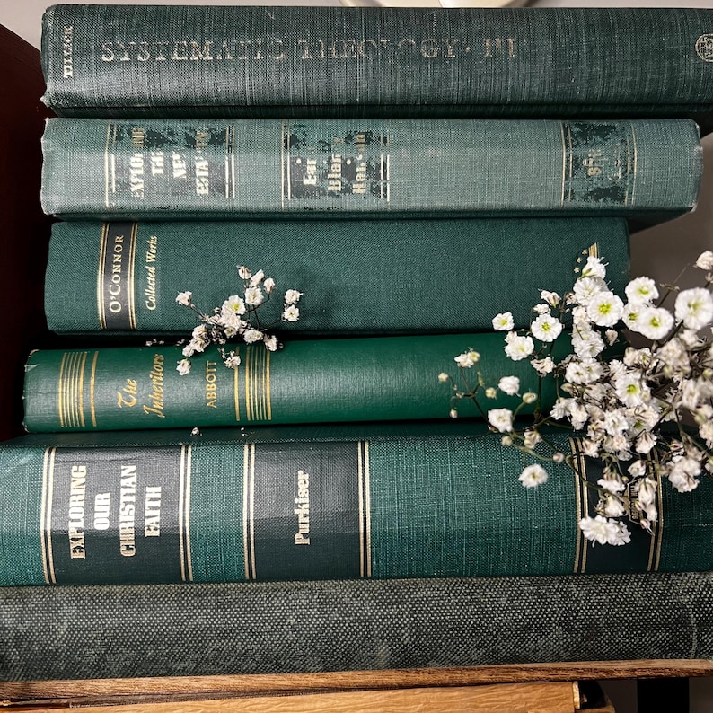 May include: A stack of vintage books in varying shades of green, with gold lettering and accents. Titles include "Systematic Theology", "Exploring Our Christian Faith", and "The Inheritors". Delicate white flowers are nestled among the books.