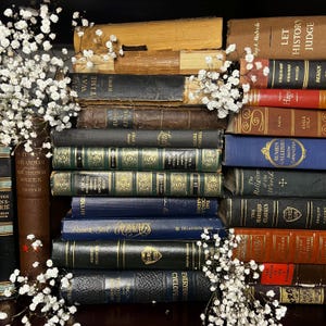 May include: A collection of antique books with various colors and sizes, stacked on a shelf. The books have aged covers and titles, including "War Time," "Grammar," and "History." White flowers are arranged around the books.