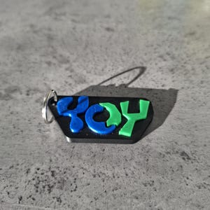 May include: Black keychain with the word "YOY" in blue and green letters. The letters are raised and have a 3D effect. The keychain has a silver ring attached.