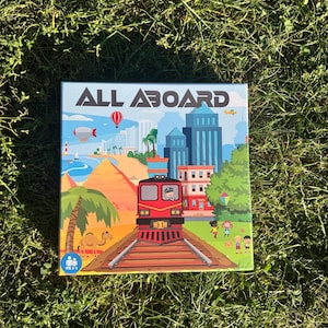 May include: A square board game box with the title "ALL ABOARD" in large, bold letters. The box art features a red train on tracks, a cityscape, and a desert scene with a camel. The background is green grass.