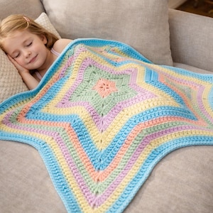 May include: A star-shaped crochet blanket featuring concentric rings of pastel colors including light blue, yellow, pink, and mint green. The handmade textile has a textured, repeating stitch pattern that radiates from the center outward to the points.