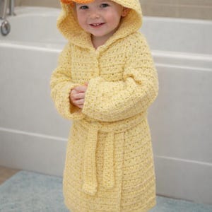 May include: A yellow crocheted bathrobe featuring a hood designed to resemble a duck, complete with black button eyes and an orange beak. The robe includes long sleeves and a matching tie belt, crafted from a textured yarn in a light yellow shade.