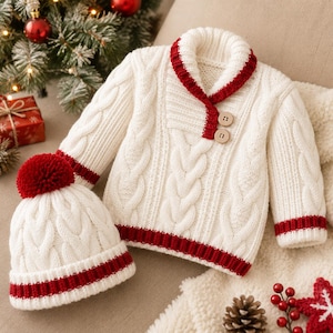 May include: A cream-coloured cable knit jumper with a shawl collar and two wooden buttons, paired with a matching cable knit beanie featuring a red pom-pom. Both items have red ribbed trim at the cuffs, hem, and collar edges.