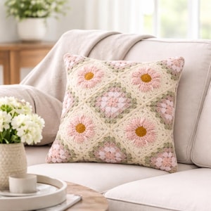 May include: A square crochet cushion featuring a repeating pattern of cream-coloured squares with pink daisy motifs and sage green accents. The design consists of alternating floral squares and smaller geometric squares, creating a textured surface.