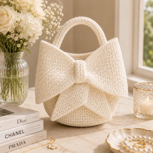 Puede incluir: Un bolso de ganchillo color crema que presenta un diseño de lazo grande y prominente en la parte frontal. El bolso tiene una base redondeada y un asa integrada, confeccionado con un patrón de punto denso y texturizado en hilo blanco roto.
