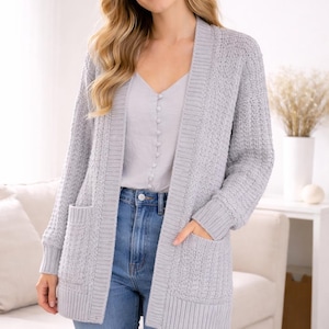 May include: A light gray open-front cardigan featuring a textured knit pattern, long sleeves with ribbed cuffs, and two front patch pockets. The garment is worn over a light gray button-down camisole and blue denim jeans, showcasing a casual layered look.