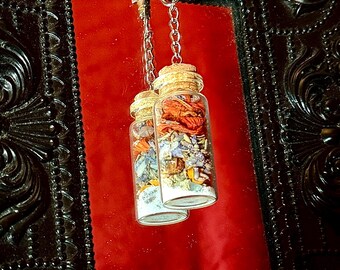May include: Two small glass bottles filled with colorful dried herbs and flowers, suspended by a silver chain. The bottles have cork stoppers and are hanging against a red background. The background is framed by a black, ornate border.