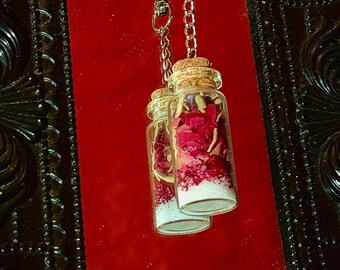 Puede incluir: Dos pequeñas botellas de vidrio llenas de pétalos de rosas rojas secas y gránulos blancos, colgadas de una cadena y un cierre plateados. Las botellas tienen tapones de corcho. El fondo es rojo vibrante, enmarcado por un borde oscuro y ornamentado.