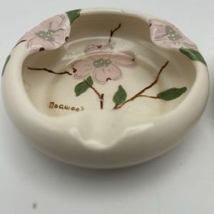 May include: Cream-coloured ceramic ashtray with a floral design. The ashtray features pink dogwood flowers, green leaves, and brown stems. The word "Dogwood" is written on the side. A classic piece for home decor.