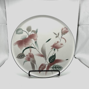 May include: A round white ceramic plate featuring a delicate floral pattern with soft pink blossoms, muted green leaves, and thin stems. The plate has a thin dark rim and is displayed on a black wire stand, showcasing a clean and simple design.
