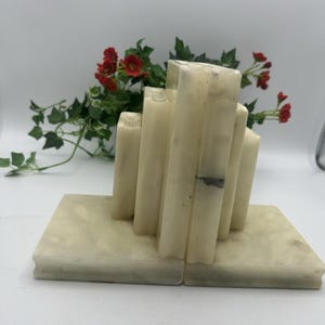 May include: A pair of off-white stone bookends featuring a stepped, tiered architectural design. Each piece consists of a flat rectangular base supporting vertical columns of varying heights, crafted from a smooth, polished natural stone material.