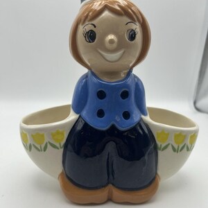 May include: A ceramic figurine of a smiling Dutch boy wearing a blue jacket and hat, with black pants. The figurine has two oval-shaped pockets decorated with yellow tulips.