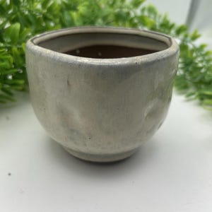 May include: A small, handmade ceramic bowl with a light gray glaze. The bowl has a rounded shape with a slightly textured surface and a darker interior. The rim is a darker shade of gray. The bowl is suitable for small plants or as a decorative item.