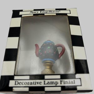 May include: A Mary Engelbreit decorative lamp finial shaped like a small black teapot with red and green accents, featuring a red handle and spout, set in a box with a black and white checkered border and the text Mary Engelbreit Decorative Lamp Finial.