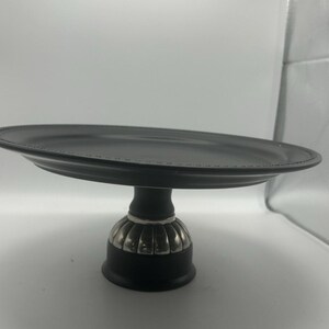 May include: A black cake stand with a round, slightly raised top and a decorative base. The base has a silver-colored, ribbed design. The stand is designed to display cakes and other desserts.