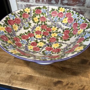 May include: A large, white ceramic serving bowl with a floral design. The bowl features a pattern of red roses, yellow flowers, and blue accents on a white background. The rim is a soft purple.