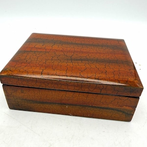 May include: A rectangular box with a glossy finish featuring a dark orange and brown crackle pattern. The lid is flat and fits securely over the base, displaying horizontal bands of color across the surface with a distinct webbed texture.