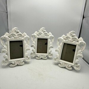 May include: Three white picture frames with ornate, swirling scrollwork detailing along the edges. Each frame features a rectangular central opening for a photograph, surrounded by a thick, decorative border with raised, curved patterns.