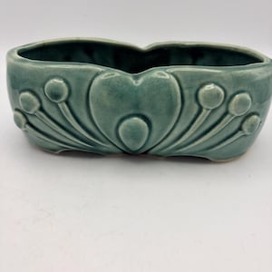May include: A sage green ceramic planter with a glossy crackle glaze finish. The exterior features a raised, symmetrical relief pattern consisting of a central heart shape flanked by radiating stems topped with small circular buds on each side.