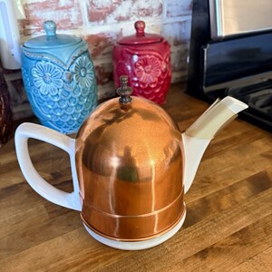 May include: A teapot featuring a white ceramic base and handle, paired with a polished copper dome-shaped cover. The lid is topped with a small, dark metal finial, and the spout is white ceramic with a metallic accent at the tip.