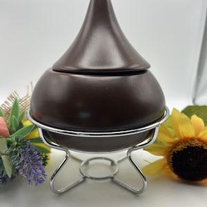 May include: A dark brown ceramic serving dish with a conical lid, resting on a chrome-colored metal stand. The dish has a rounded base and a smooth, glossy finish. The stand has a circular base and four curved legs.