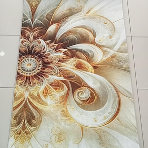 Op de afbeelding: Een rechthoekig vloerkleed met een groot, centraal abstract bloemmotief in goud-, brons-, bruin- en cr&egrave;metinten. Het patroon bestaat uit wervelende, gelaagde bloemblaadjes en complexe, vloeiende lijnen die vanuit het midden naar buiten stralen.