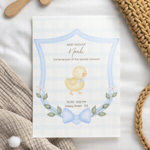 Puede incluir: Una invitaci&oacute;n para baby shower con una ilustraci&oacute;n en acuarela de un patito amarillo con una pajarita azul. La invitaci&oacute;n tiene un marco azul en forma de escudo con detalles florales y el nombre "Noah". El texto "Arte que inspira infinitas posibilidades" est&aacute; en la parte superior.