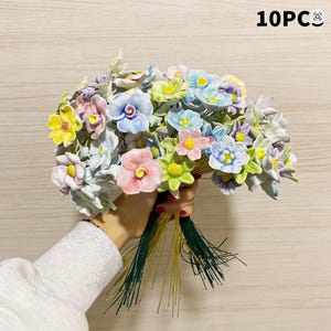 以下が含まれることがあります： ピンク、黄色、青、緑のパステルカラーのセラミックフラワーブーケ。 花は緑色の茎でまとめられており、「10PC」の文字が見えます。 花は繊細で手作りの外観をしています。