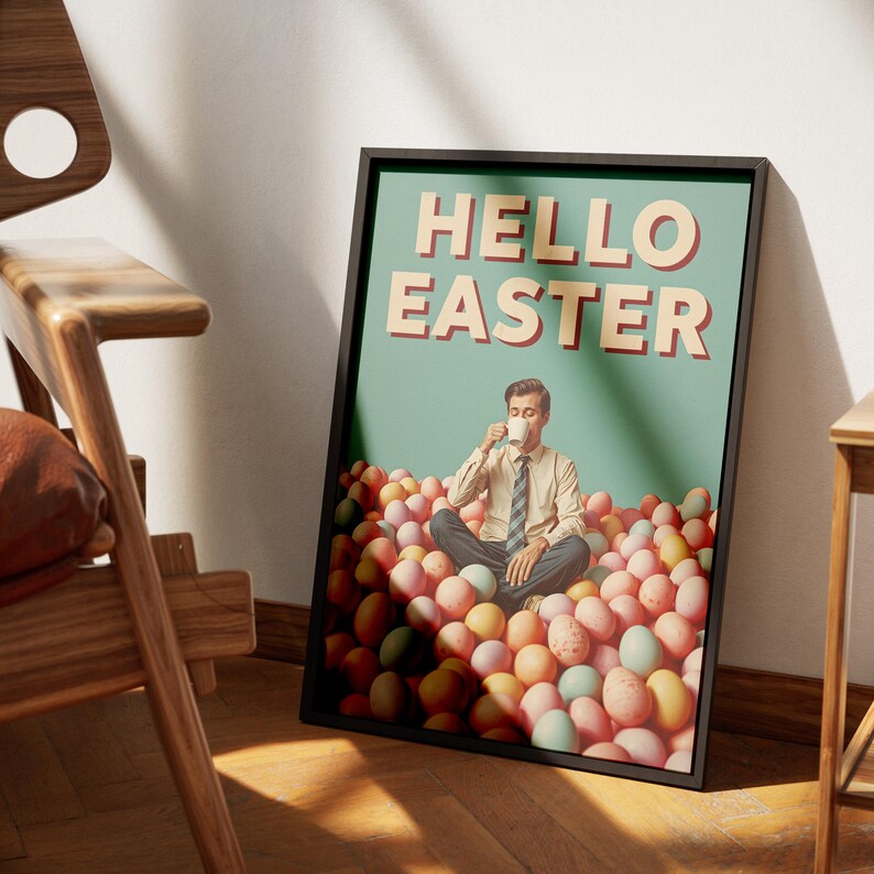 May include: A framed poster with the text "HELLO EASTER" in a retro font. The poster shows a man seated in a pile of pastel-coloured Easter eggs, drinking from a mug. The poster is in a black frame and leans against a wall.