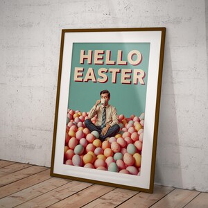 May include: Framed poster with the words "HELLO EASTER" in red and white. A man is seated in a pile of pastel-coloured Easter eggs, holding a cup. The poster is in a brown frame, leaning against a white wall and wooden floor.