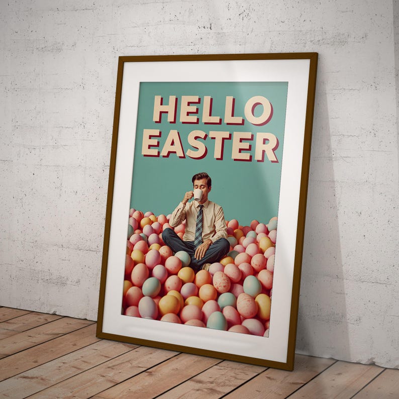 May include: Framed poster with the words "HELLO EASTER" in red and white. A man is seated in a pile of pastel-coloured Easter eggs, holding a cup. The poster is in a brown frame, leaning against a white wall and wooden floor.