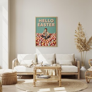 May include: A framed art print with the words "HELLO EASTER" in a retro font. The artwork depicts a person sitting in a pile of colourful Easter eggs, holding a cup. The print is displayed in a living room setting with a sofa and coffee table.