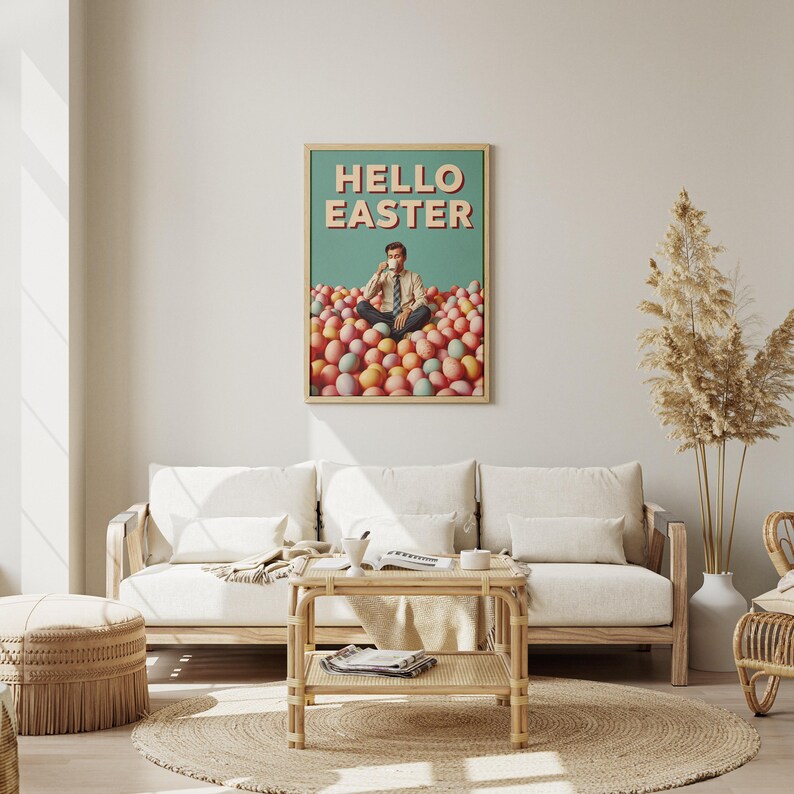 May include: A framed art print with the words "HELLO EASTER" in a retro font. The artwork depicts a person sitting in a pile of colourful Easter eggs, holding a cup. The print is displayed in a living room setting with a sofa and coffee table.