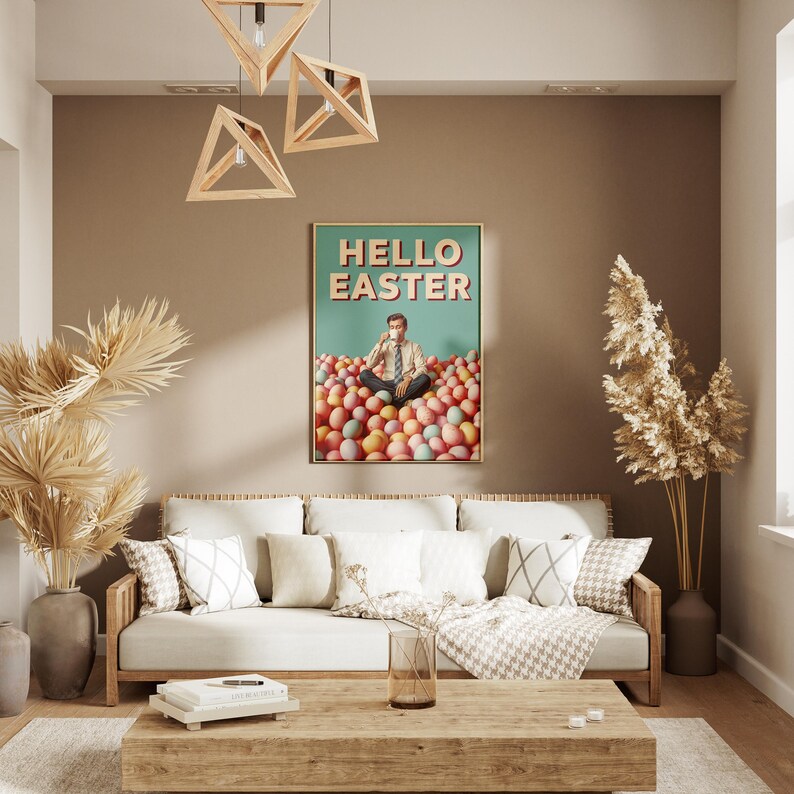 May include: A living room interior with a framed poster saying "HELLO EASTER". The poster shows a person sitting in a pile of Easter eggs. A light-coloured sofa with cushions and a wooden coffee table are in front of the poster. Wooden light fixtures hang from the ceiling.