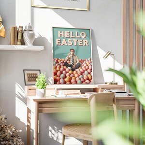 May include: A framed print with the words "HELLO EASTER" on a teal background. The image inside the frame depicts a person sitting in a pile of Easter eggs. The print is displayed on a wooden desk with a small lamp and a chair.